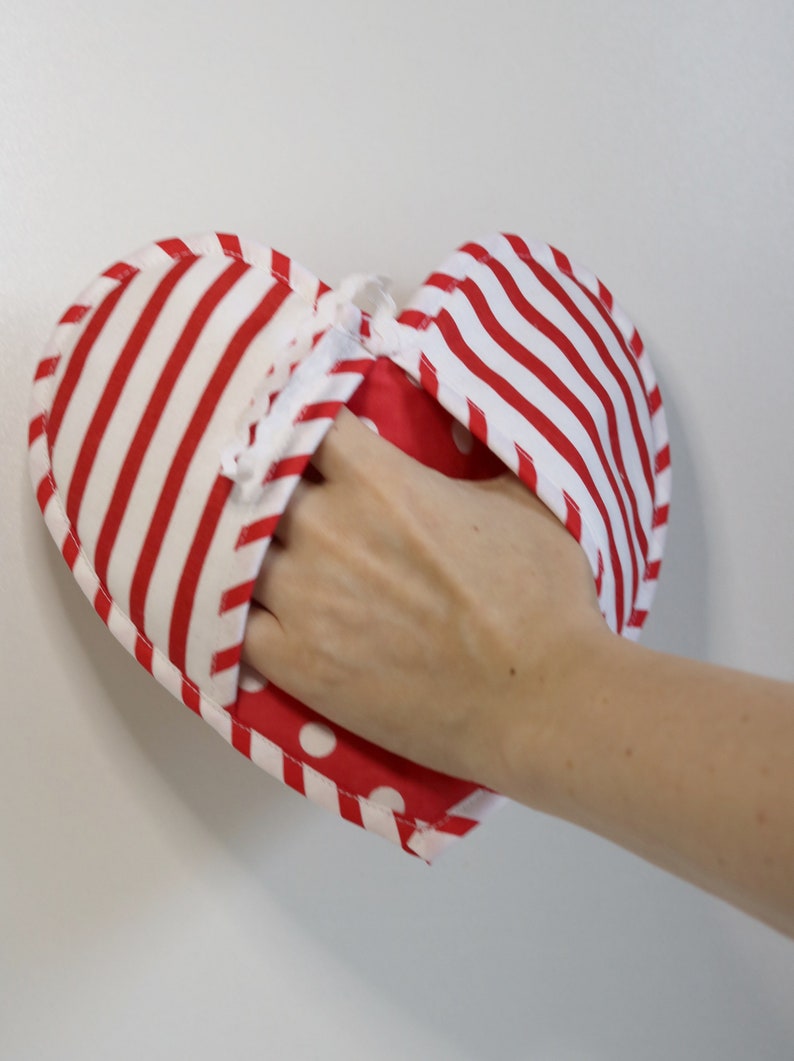 May include: A red and white striped heart-shaped pot holder with white polka dots on the inside. The pot holder is made of fabric and has a white string attached to it.