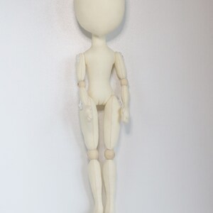 May include: A white fabric doll with wooden joints. The doll has a round head and no facial features. The doll is standing upright with its arms at its sides.