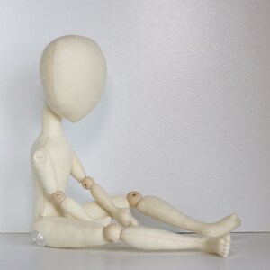 May include: A white fabric doll with wooden joints, sitting with its legs crossed. The doll has a simple, round head and no facial features.