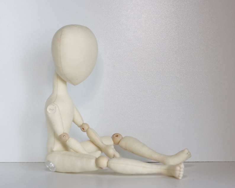 May include: A white fabric doll with wooden joints, sitting with its legs crossed. The doll has a simple, round head and no facial features.