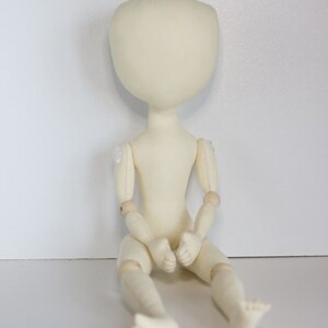 May include: A white fabric doll with wooden joints, sitting with its legs crossed. The doll has a blank face and is ready to be customized.