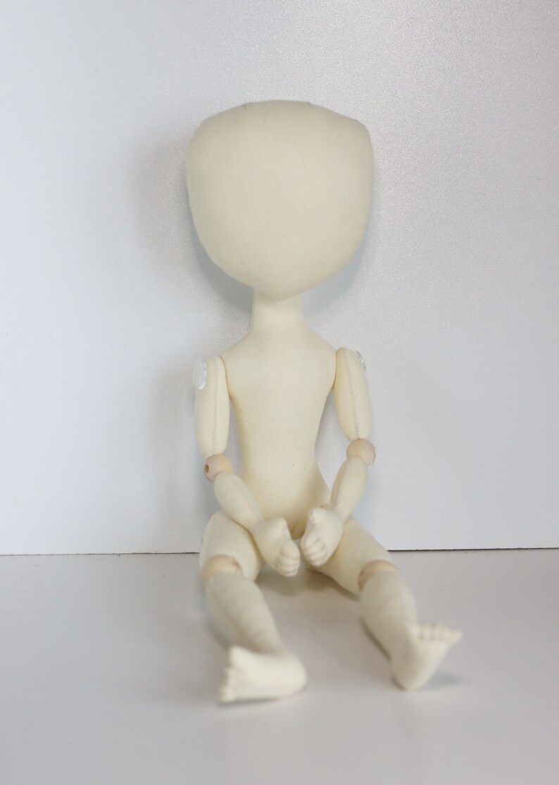 May include: A white fabric doll with wooden joints, sitting with its legs crossed. The doll has a blank face and is ready to be customized.