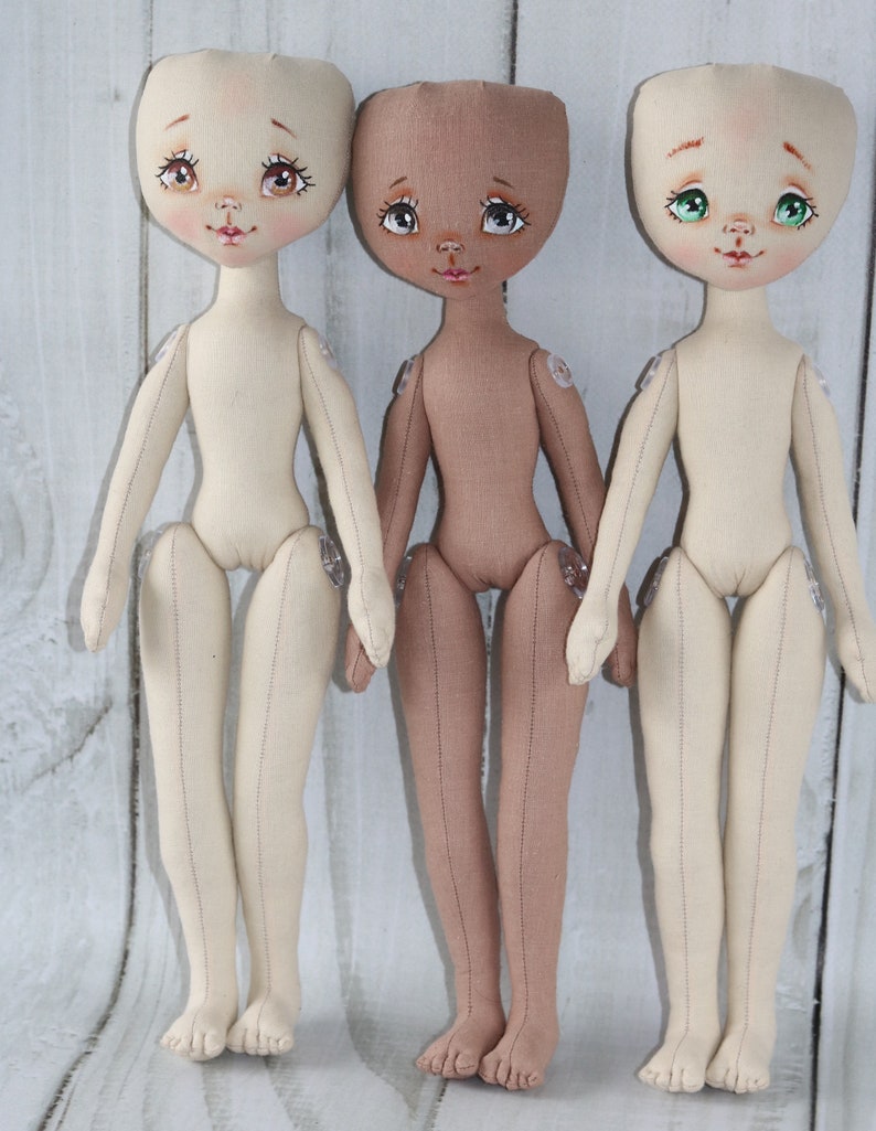 May include: Three handmade fabric dolls with different skin tones. The dolls have painted faces with brown eyes, pink lips, and rosy cheeks. The dolls are standing on a white wooden surface.