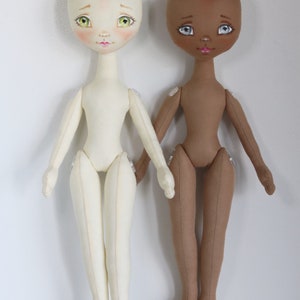 May include: Two fabric dolls with painted faces and sewn details. One doll has a light skin tone and the other has a brown skin tone. Both dolls have green eyes and pink lips.