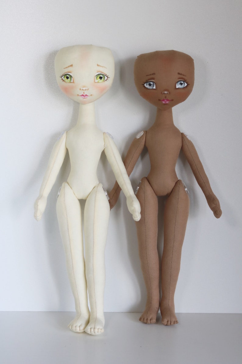 May include: Two fabric dolls with painted faces and sewn details. One doll has a light skin tone and the other has a brown skin tone. Both dolls have green eyes and pink lips.