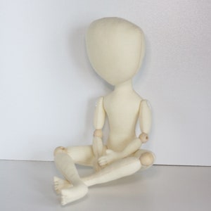 May include: A white fabric doll with wooden joints, sitting with its legs crossed. The doll has a simple, round head and no facial features.