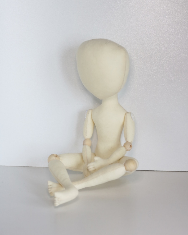 May include: A white fabric doll with wooden joints, sitting with its legs crossed. The doll has a simple, round head and no facial features.