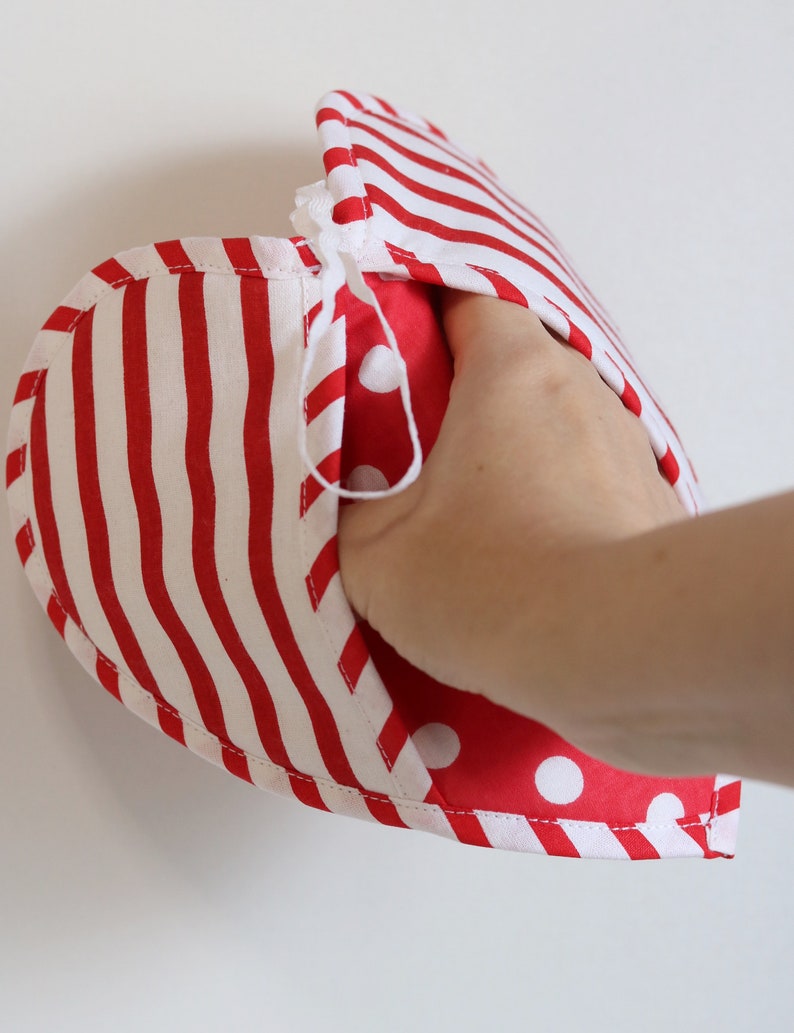 May include: Red and white striped oven mitt with a red and white polka dot lining. The mitt is folded in half and a hand is holding it open.