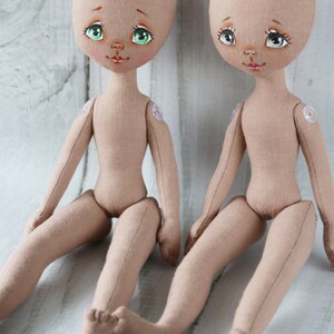 May include: Two unpainted fabric dolls with green eyes and painted facial features are sitting on a white wooden surface. The dolls have button eyes and are made of a beige fabric.