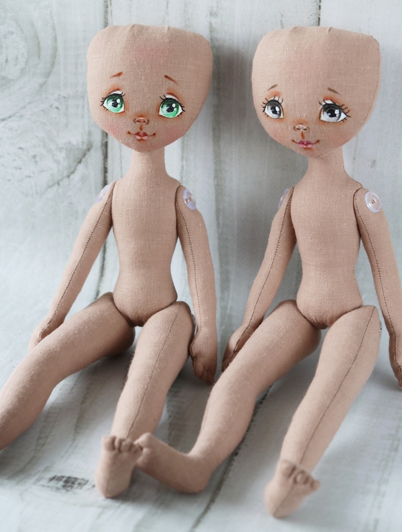 May include: Two unpainted fabric dolls with green eyes and painted facial features are sitting on a white wooden surface. The dolls have button eyes and are made of a beige fabric.