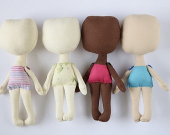 Op de afbeelding: Vier handgemaakte stoffen poppen met verschillende huidtinten en outfits. De poppen staan in een rij op een witte achtergrond. Elke pop heeft een eenvoudige, gestikte outfit. De poppen kijken allemaal naar voren.