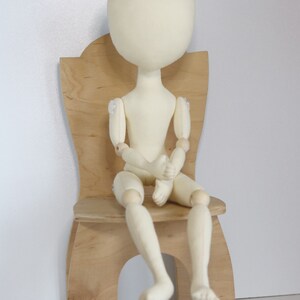 May include: A white fabric doll with wooden joints sits on a wooden chair. The doll has a simple, round head and no facial features.