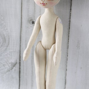 May include: A plain white fabric doll with blue eyes and a stitched face. The doll has a simple design with no clothing or accessories.