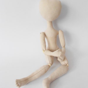 May include: A beige fabric doll with jointed limbs, sitting with its arms crossed. The doll has a simple, round head with no facial features.