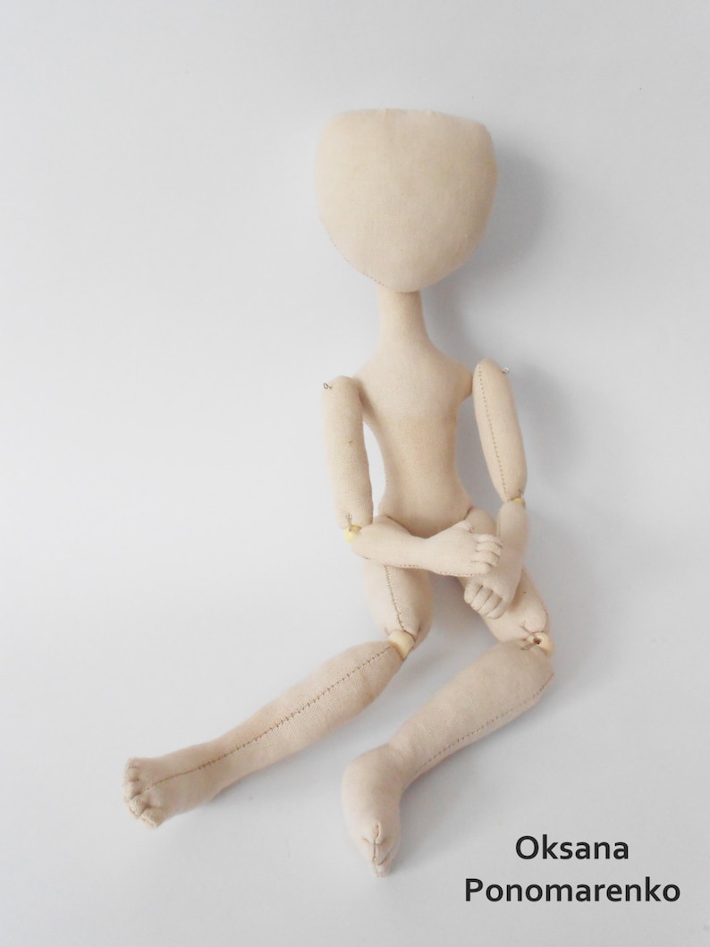 May include: A beige fabric doll with jointed limbs, sitting with its arms crossed. The doll has a simple, round head with no facial features.