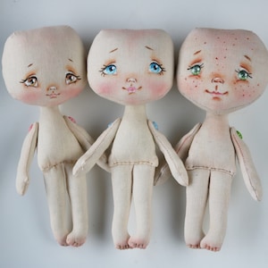 May include: Three handmade fabric dolls with painted faces. The dolls have brown, blue, and green eyes, and rosy cheeks. They are all wearing simple white fabric clothing.