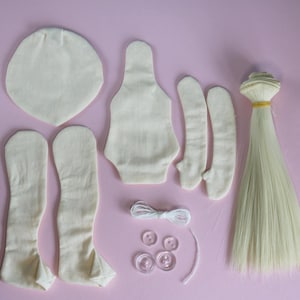 May include: A doll-making kit with various fabric pieces in cream, including a head, body, arms, and legs. Also included are a bundle of blonde hair, clear buttons, white thread, and a needle. The items are arranged on a pink surface.
