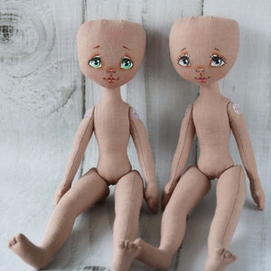 May include: Two unpainted fabric dolls with green and brown eyes, painted lips, and sewn-on details. The dolls are sitting on a white wooden surface.