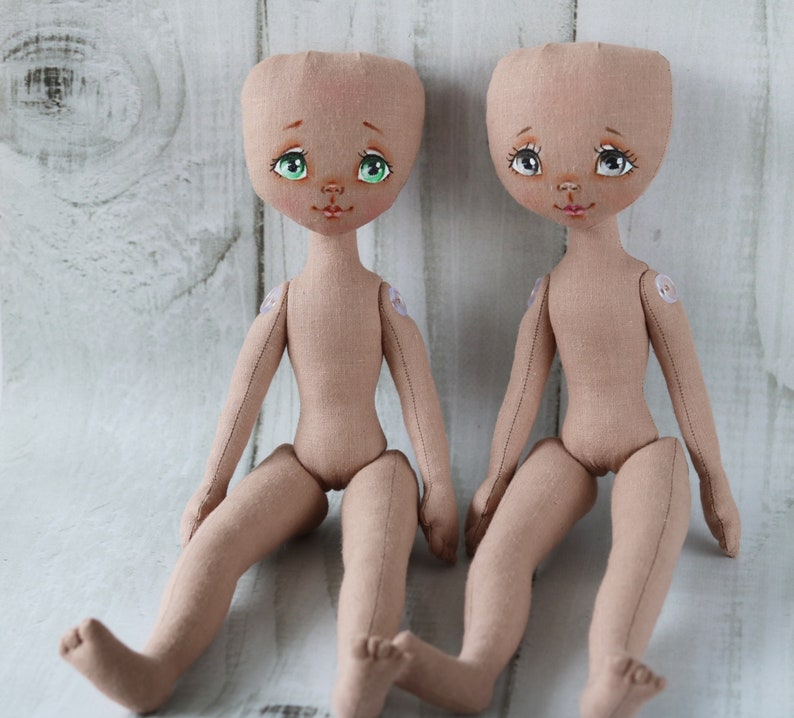 May include: Two unpainted fabric dolls with green and brown eyes, painted lips, and sewn-on details. The dolls are sitting on a white wooden surface.