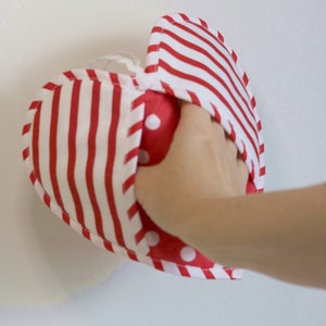 May include: A red and white striped heart-shaped fabric pouch with red polka dots on the inside. The pouch is being held open by a hand.