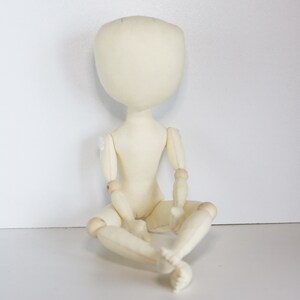 May include: A white fabric doll with wooden joints, sitting with its legs crossed. The doll has a blank face and no hair.