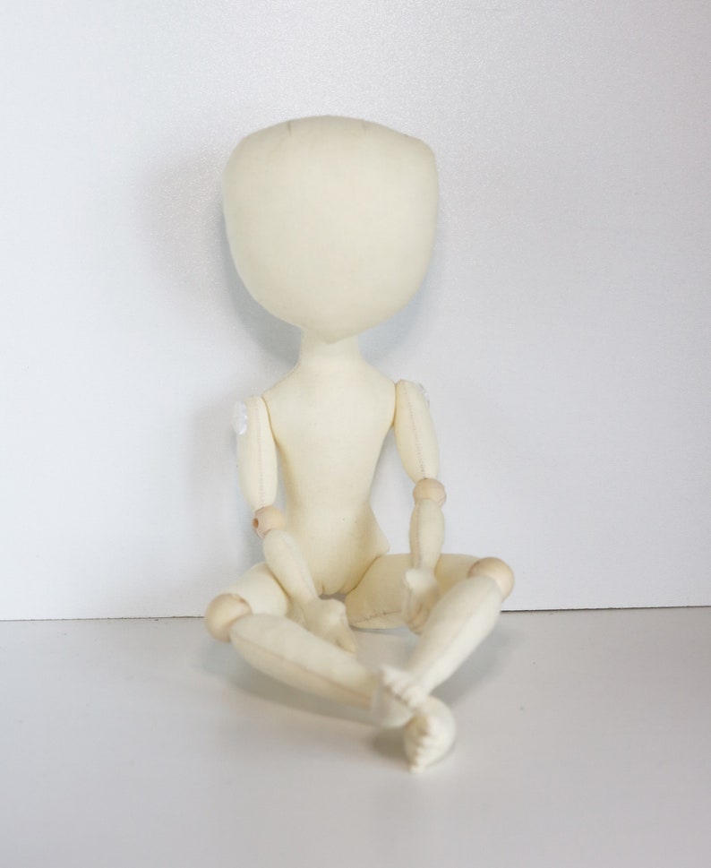 May include: A white fabric doll with wooden joints, sitting with its legs crossed. The doll has a blank face and no hair.
