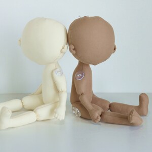 May include: Two fabric dolls sitting side-by-side. The doll on the left is beige and the doll on the right is brown. Both dolls have a simple design with no facial features.