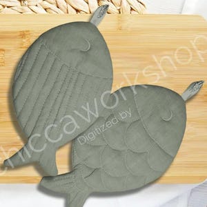 Può includere: Due presine grigie a forma di pesce su un tagliere di legno. Una presina ha cuciture verticali, l'altra ha cuciture a forma di scaglia. Le presine hanno pinne e un piccolo anello nella parte superiore. Lo sfondo comprende un tappeto intrecciato e un panno bianco.