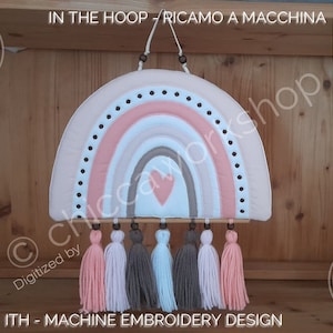 May include: A pink, white and brown rainbow with a heart in the centre. The rainbow is hanging from a wooden dowel with tassels in pink, white and brown. The image includes the text "IN THE HOOP - RICAMO A MACCHINA" and "ITH - MACHINE EMBROIDERY DESIGN".