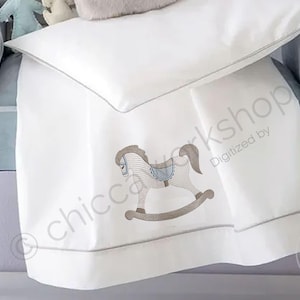 May include: White baby bedding set with a rocking horse embroidery. The set includes a pillow and a blanket with a gray trim. The rocking horse is in shades of brown and blue. The bedding is on a light blue upholstered chair.