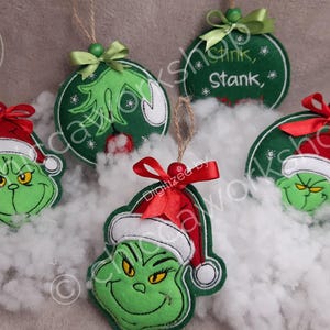 Puede incluir: Una colección de adornos navideños verdes con la cara del Grinch y otros diseños festivos. Cada adorno tiene un lazo rojo o verde y una cuerda para colgar. Algunos adornos tienen el texto "Stink, Stank, Stunk".