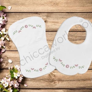 May include: White baby bib and burp cloth set with embroidered pink flowers and green leaves. The bib has a curved shape with a neck opening. The burp cloth is oval. Both are on a wooden background.