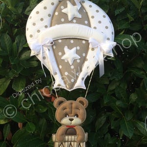 May include: A decorative hot air balloon with a teddy bear in a basket. The balloon is white with grey polka dots and star accents. The teddy bear is brown with a red collar. White ribbons and bows add detail.