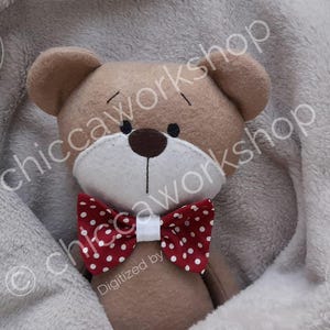 Könnte beinhalten: Ein brauner Teddybär mit weißem Gesicht und einer rot-weißen gepunkteten Schleife. Der Bär liegt auf einer grauen Decke.