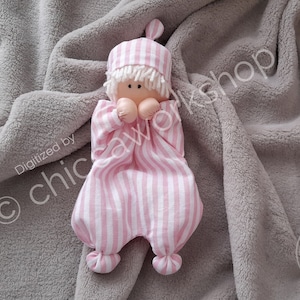 以下が含まれることがあります： ピンクと白のストライプの布製のドールで、白い毛糸の髪とピンクと白のストライプの帽子をかぶっています。ドールは灰色のふわふわの毛布の上に横たわっています。