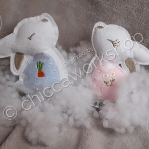 May include: Two plush bunny toys, one blue and one pink, with embroidered details. The blue bunny has a carrot on its shirt and the pink bunny has flowers. The bunnies are sitting on a white fluffy surface.