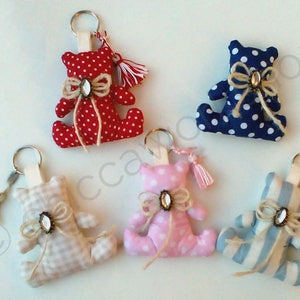 Può includere: Cinque portachiavi a forma di orsetto fatti a mano in vari modelli e colori. Gli orsetti sono rossi a pois bianchi, blu a pois bianchi, rosa a pois bianchi, a quadretti beige e a righe blu e bianche. Ogni orsetto ha un fiocco e una gemma decorativa.