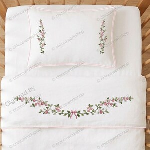 Può includere: Un set di biancheria da culla bianco composto da una federa e una coperta con delicati ricami floreali. La federa presenta due steli floreali verticali, mentre la coperta mostra una ghirlanda floreale centrale con un piccolo fiocco rosa.