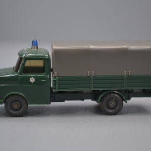 以下が含まれることがあります： グレーのキャンバスの幌が荷台を覆っている緑の玩具トラック。トラックの上には青いライトがあり、側面には白い星があります。