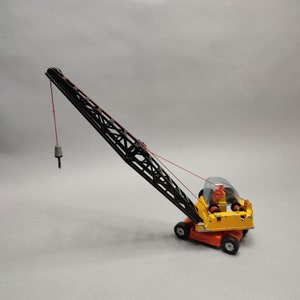 May include: A yellow and black toy crane with a red cable and a gray hook. The crane is on a white background.