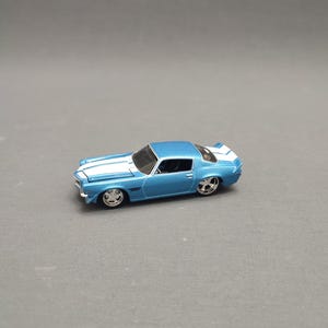 Può includere: Un modello di auto giocattolo blu e bianco di una Chevrolet Camaro classica. L'auto ha strisce da corsa bianche e dettagli neri.