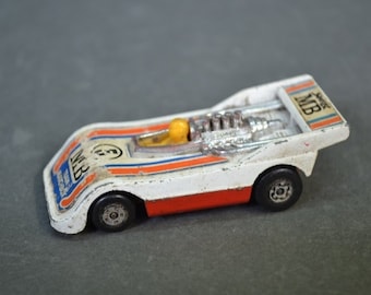 May include: White and orange toy race car with a silver engine and a yellow driver. The car has the text "MB" on the side.