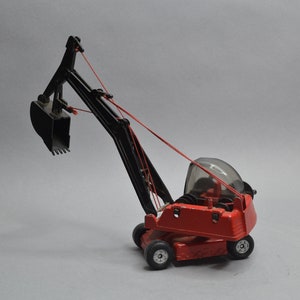 May include: A red toy construction vehicle with a black extendable arm and a black bucket. The vehicle has a clear plastic cab and black wheels.