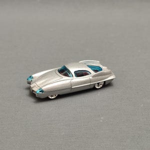 Könnte beinhalten: Ein silbernes Spielzeugauto mit einer blauen Windschutzscheibe. Das Auto hat ein abgerundetes Design und ist ein Coupé.