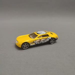 Puede incluir: Coche de juguete amarillo con diseños gráficos en blanco y negro en el lateral y el techo. El coche tiene ruedas negras y un interior beige. Es una réplica en miniatura de un coche deportivo.