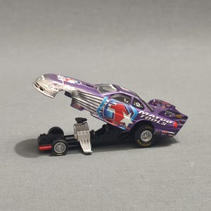 Può includere: Modello in scala 1/64 di un dragster in metallo pressofuso, di colore viola e argento, con logo Matco Tools. L'auto è in posizione di impennata, con le ruote anteriori sollevate da terra.