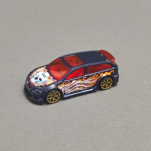 Puede incluir: Un coche de juguete azul con un diseño de calavera y llamas. El coche tiene un techo rojo y un parabrisas transparente. El coche es un juguete Hot Wheels.