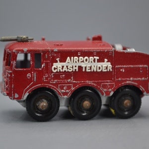 May include: Red toy fire truck with a white "Airport Crash Tender" label. The truck has a ladder on top and four wheels.