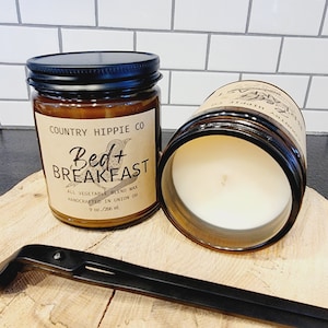 May include: Two glass jars of scented candles with black lids. The labels read "Country Hippie Co Bed + Breakfast" and "All Vegetable Blend Wax". Each candle is 9 oz. The jars are on a wooden surface.