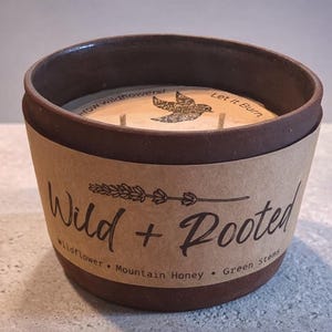 May include: A brown ceramic candle holder with a paper label that reads "Wild + Rooted." The candle has a wooden top with the text "Let It Burn" and a bird graphic. The scent is wildflower, mountain honey, and green stems.